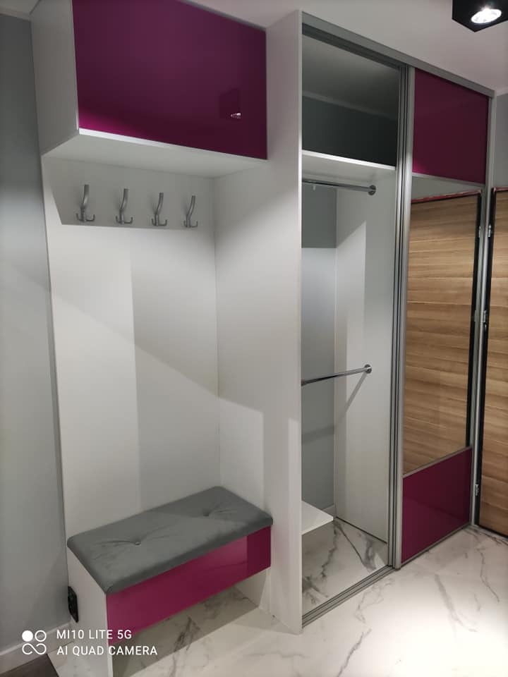 Biała szafa wnękowa z siedziskiem i wieszakami, akcenty w kolorze fuksji, przesuwne drzwi z lustrem i drewnianym panelem, marmurowa podłoga.