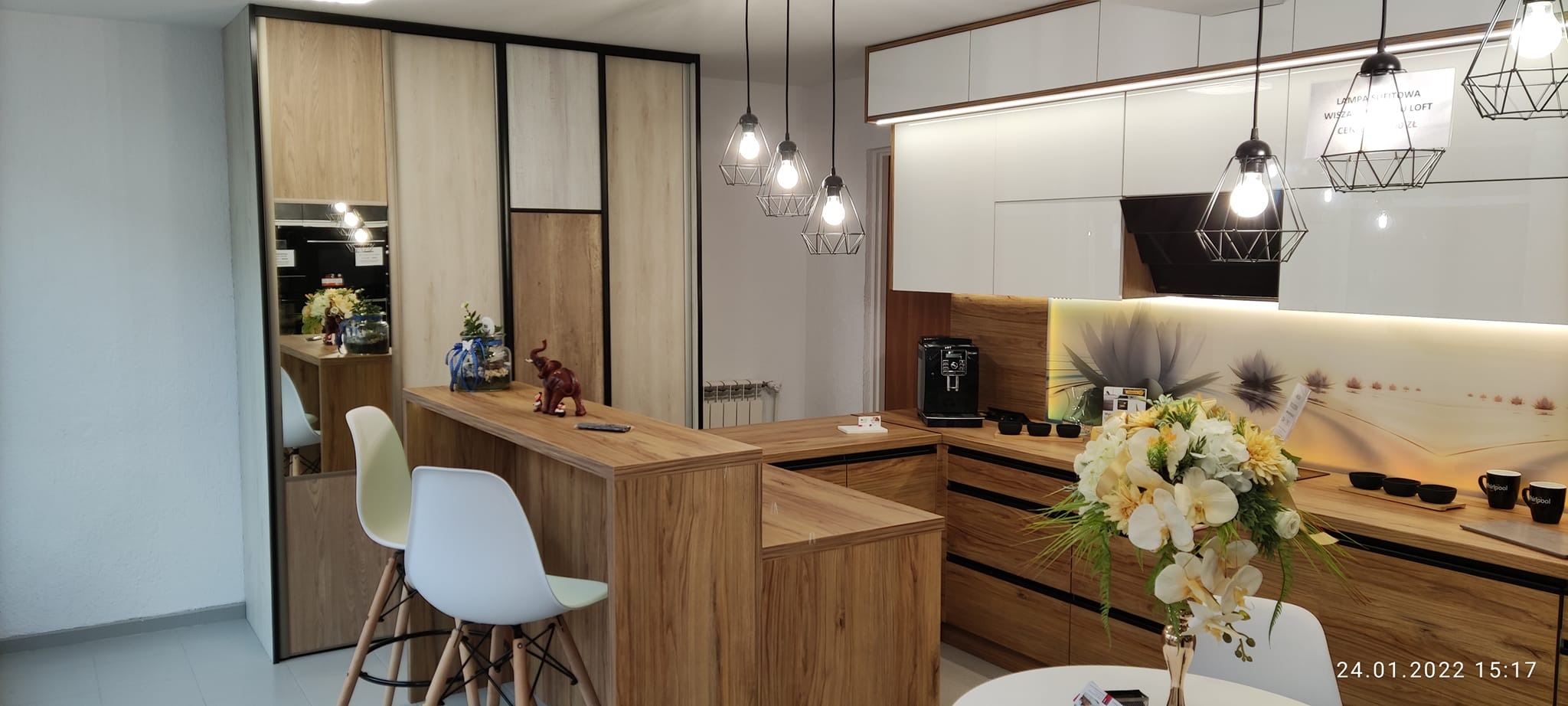 Nowoczesna kuchnia z białymi szafkami górnymi, drewnianymi szafkami dolnymi i blatem, wyspą z hokerami, oświetleniem wiszącym w stylu loft i dekoracyjnym panelem ściennym z motywem kwiatowym.