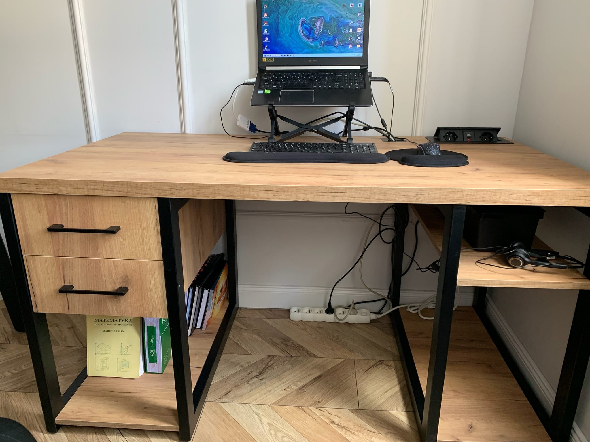Biurko z blatem drewnopodobnym i czarnymi metalowymi nogami, z szufladami po lewej stronie i półkami po prawej, na blacie laptop na podstawce, klawiatura, myszka i wbudowane gniazdka elektryczne...