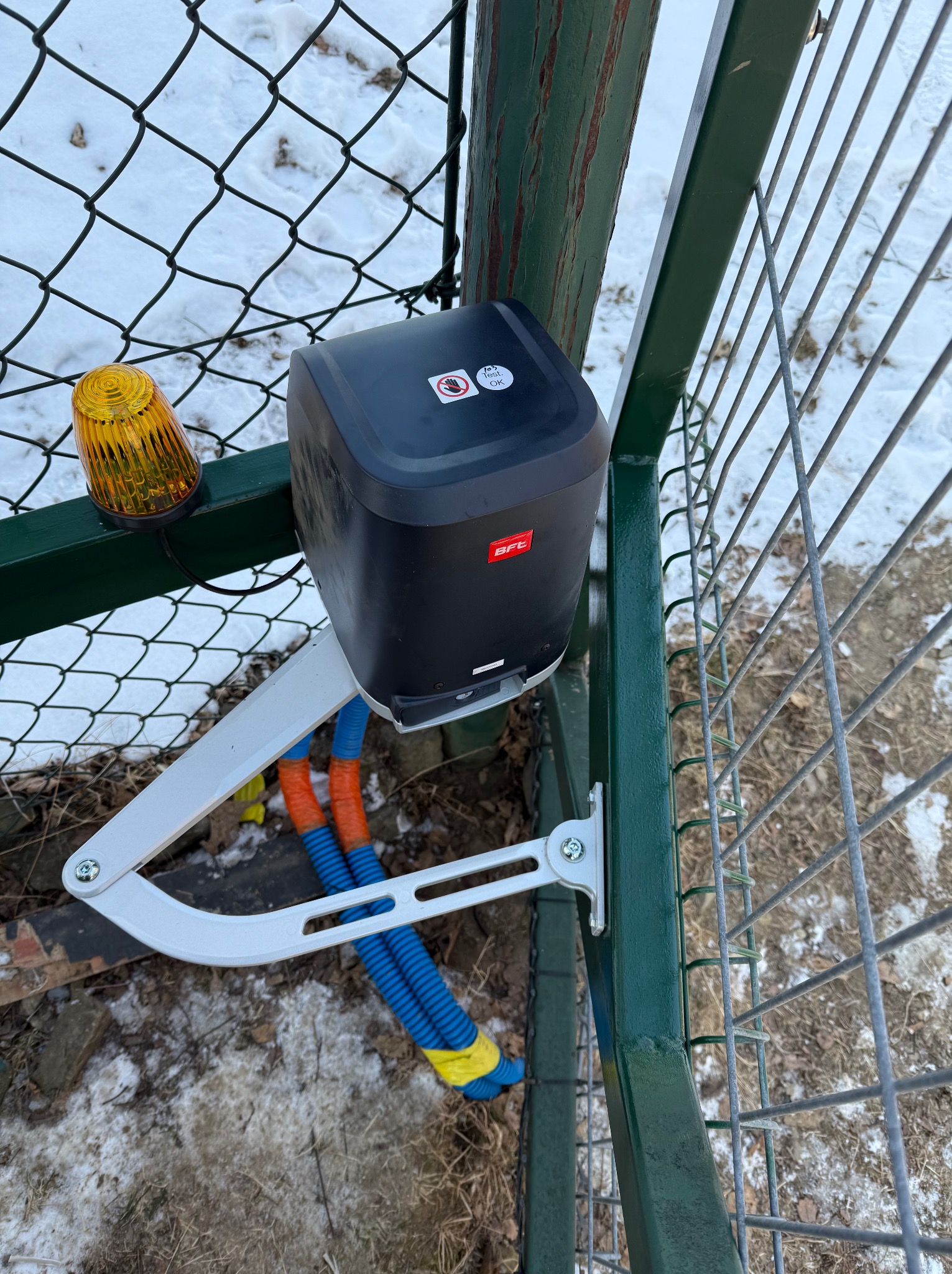 Zamontowany napęd BFT z pomarańczową lampą ostrzegawczą na metalowej bramie posesji, widoczne przewody w peszlach. Zima.