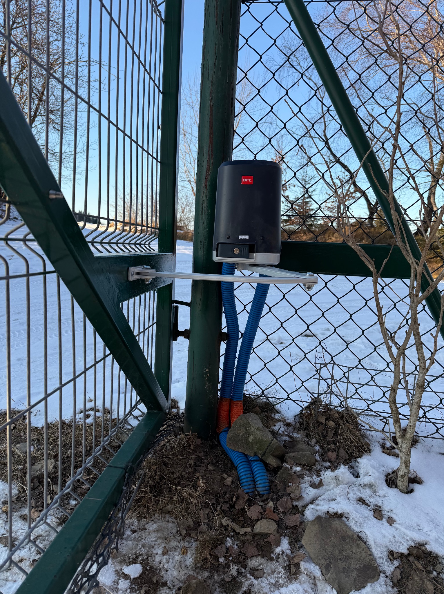 Zamontowany napęd BFT do bramy posesyjnej z widocznymi niebieskimi przewodami w peszlach, umieszczony na tle siatki ogrodzeniowej i zimowego krajobrazu.