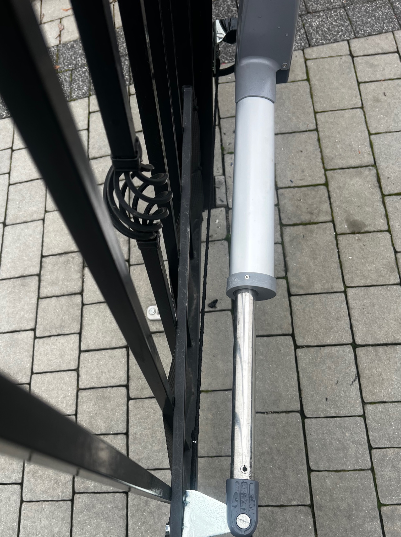 Zamknięta, czarna, metalowa brama z zamontowanym siłownikiem automatyki na tle szarej kostki brukowej. Widok z góry, zbliżenie na mechanizm.