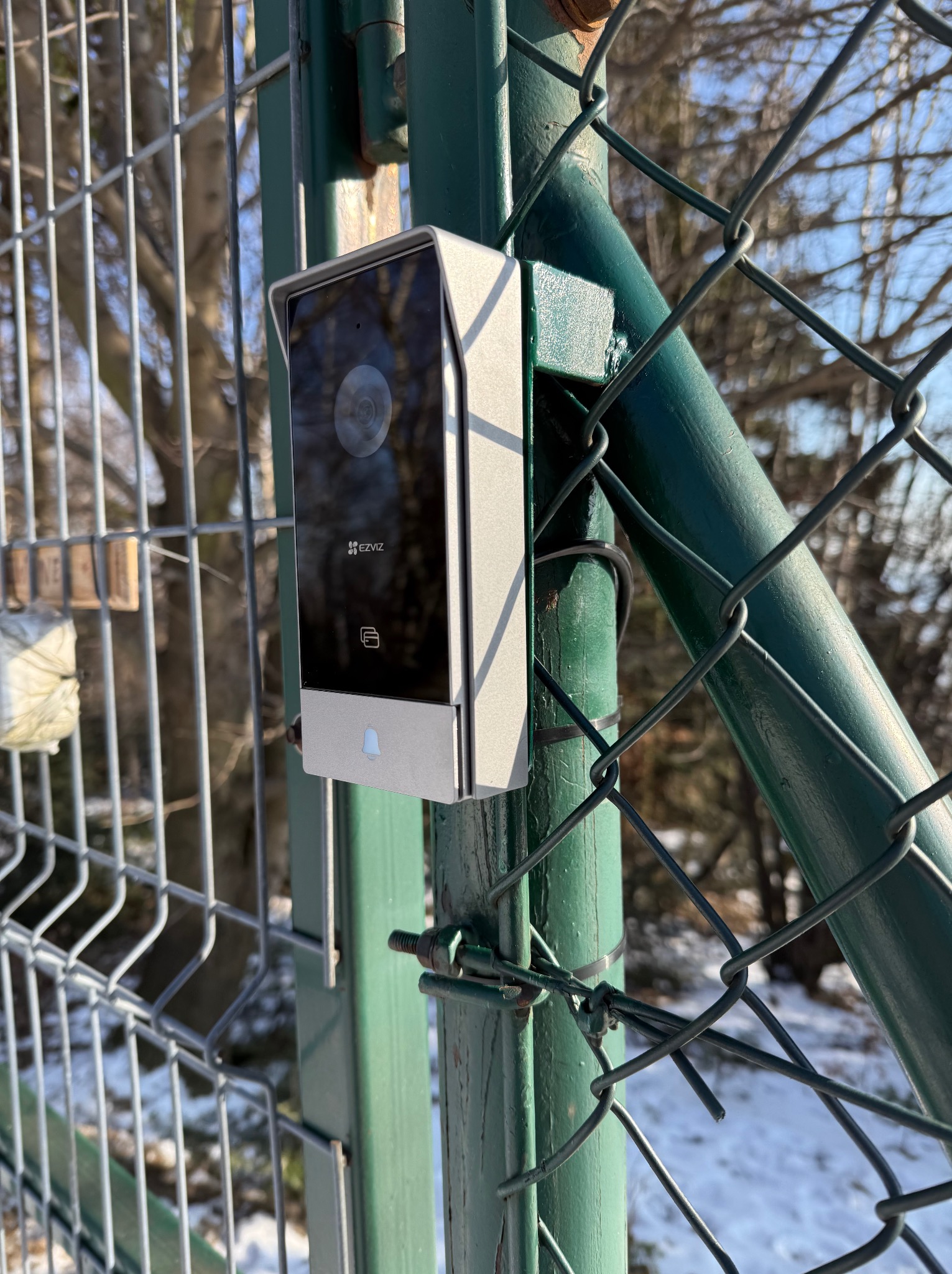 Zamontowany wideodomofon marki EZVIZ na zielonym słupku ogrodzenia z siatki. Widoczne elementy montażowe i fragment zimowego krajobrazu w tle.