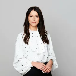 Portret kobiety w białej bluzce w czarne kropki i czarnej spódnicy, z rękami złożonymi przed sobą, z delikatnym uśmiechem i pierścionkiem na palcu.