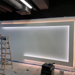 Ściana z podświetleniem LED w trakcie prac wykończeniowych, widoczne drabiny malarskie, taśmy zabezpieczające, oraz sprzęt do szlifowania gładzi.