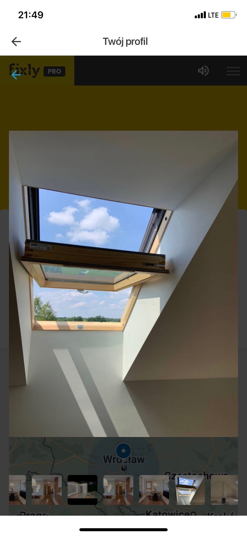 Otwarty drewniany okno dachowe z widokiem na błękitne niebo z chmurami i zielenią, białe ściany poddasza z widocznymi promieniami słonecznymi.