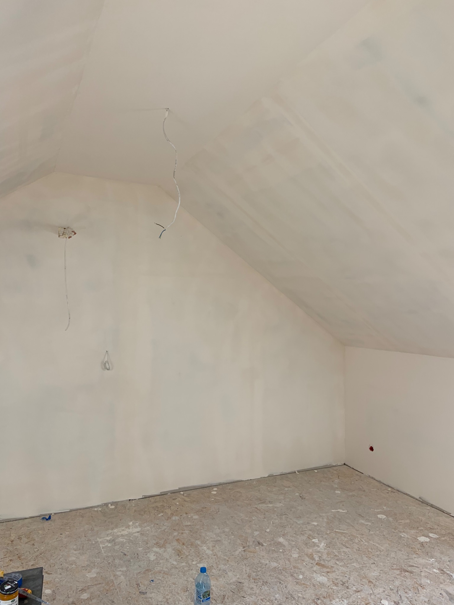 Poddasze po nałożeniu gładzi na ściany i sufit, widoczne kable elektryczne, podłoga zabezpieczona płytami OSB, butelka wody i narzędzia malarskie w rogu pomieszczenia.