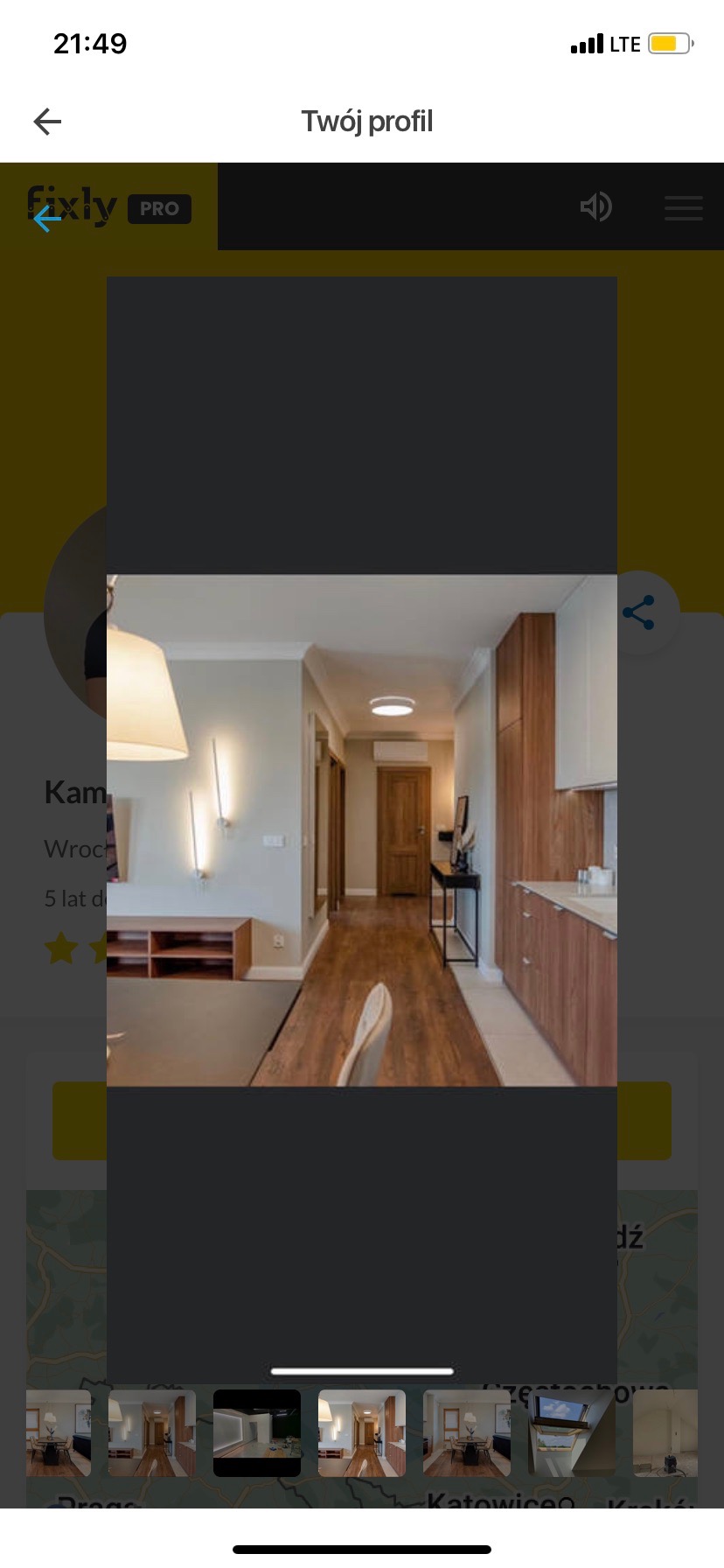 Wnętrze mieszkania po remoncie: widok na przedpokój z drewnianą podłogą, szafkami kuchennymi i oświetleniem ściennym. Częściowo widoczny stół z krzesłem.