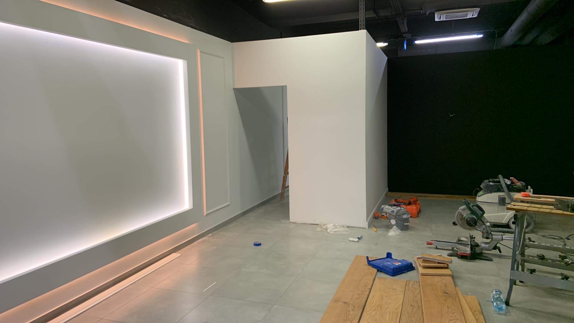 Wnętrze biura w trakcie prac wykończeniowych: ściany pomalowane na szaro z dekoracyjnym oświetleniem LED, widoczne narzędzia budowlane i deski podłogowe.