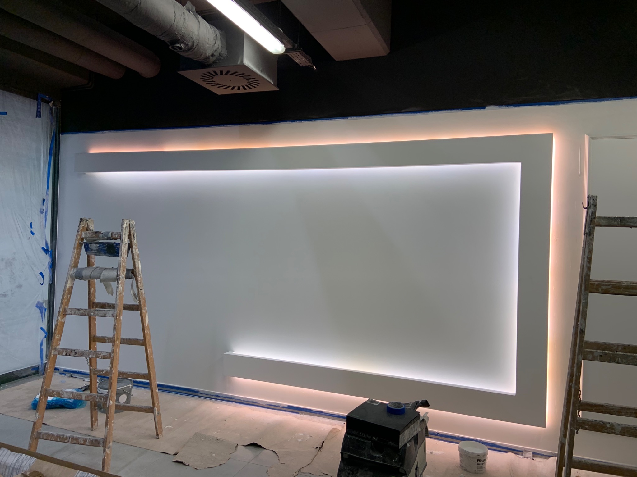 Ściana z podświetleniem LED w trakcie prac wykończeniowych, widoczne drabiny malarskie, taśmy zabezpieczające, oraz sprzęt do szlifowania gładzi.