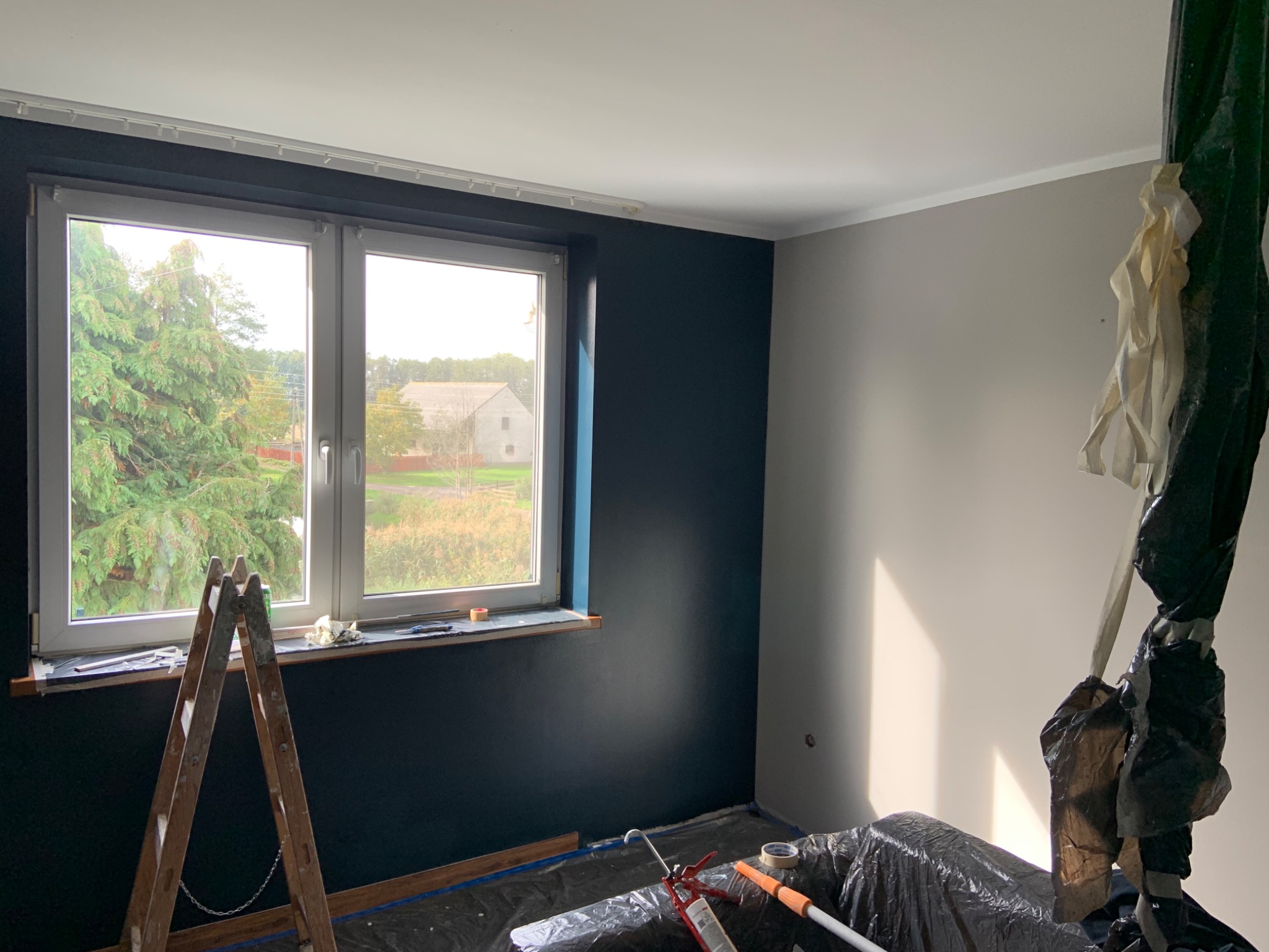 Pokój po częściowym pomalowaniu: jedna ściana ciemnogranatowa, druga jasnoszara, okno z widokiem na zieleń, drabina oparta o ścianę, podłoga zabezpieczona folią malarską.