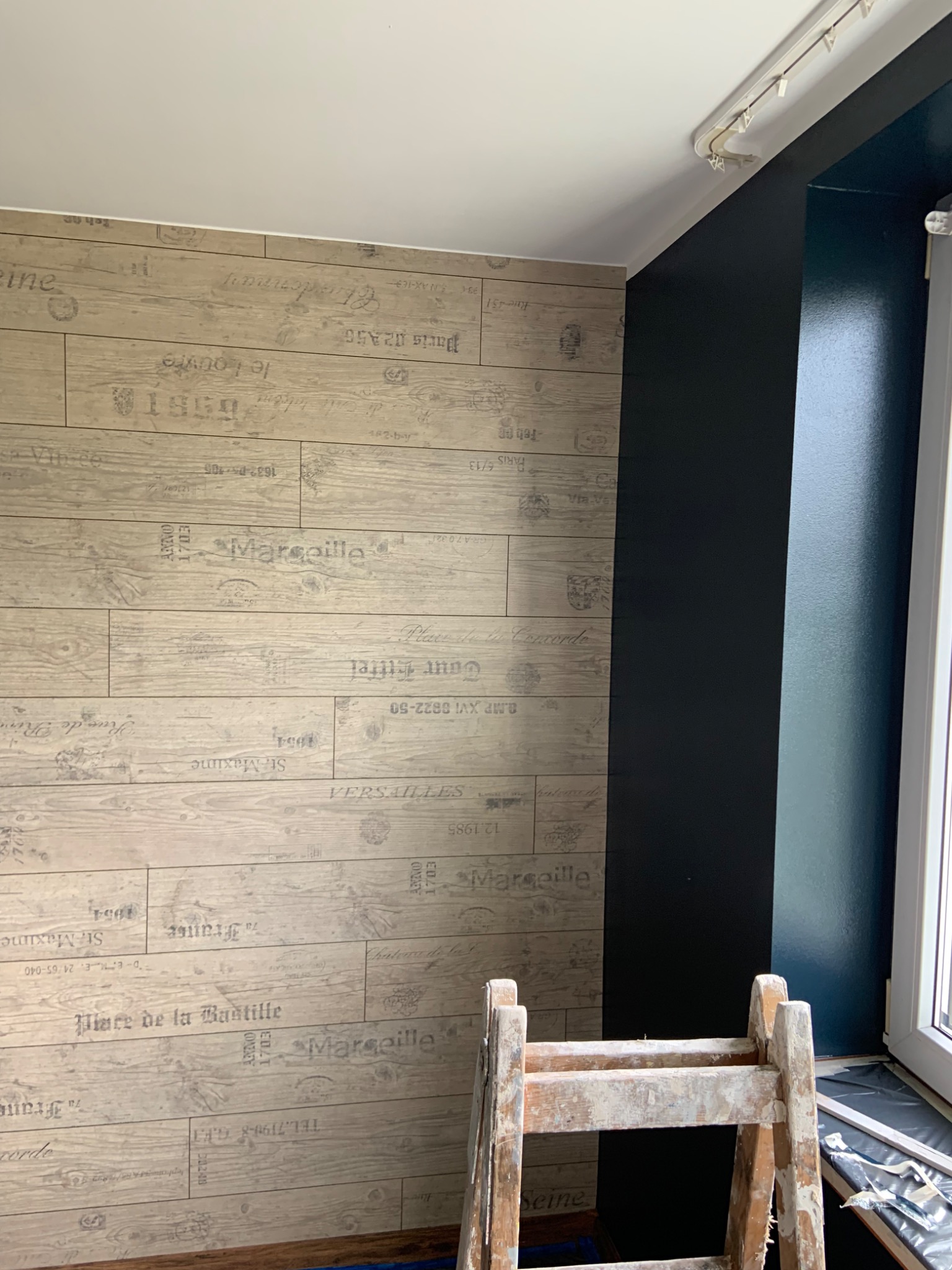 Ściana z tapetą imitującą drewniane panele z nadrukowanymi napisami w stylu retro, sąsiadująca z ciemną ścianą przy oknie, widoczny fragment drabiny malarskiej.