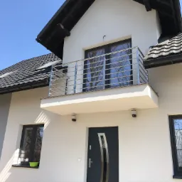 Biały dom z balkonem i balustradą ze stali nierdzewnej, widok z dołu na tle błękitnego nieba.