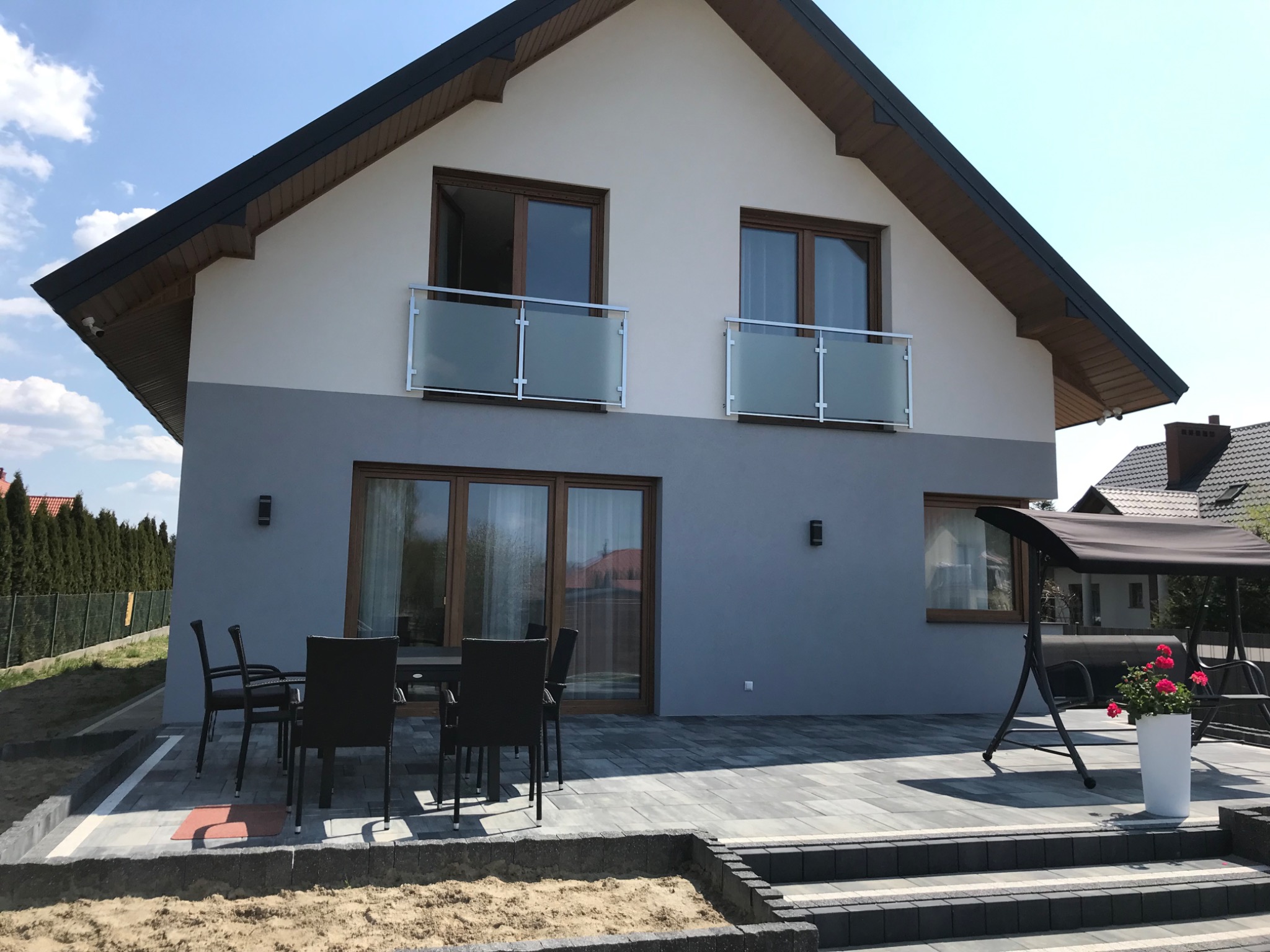 Nowoczesny dom dwukondygnacyjny z balkonami zabezpieczonymi szkłem, widok z zewnątrz na taras z meblami ogrodowymi.