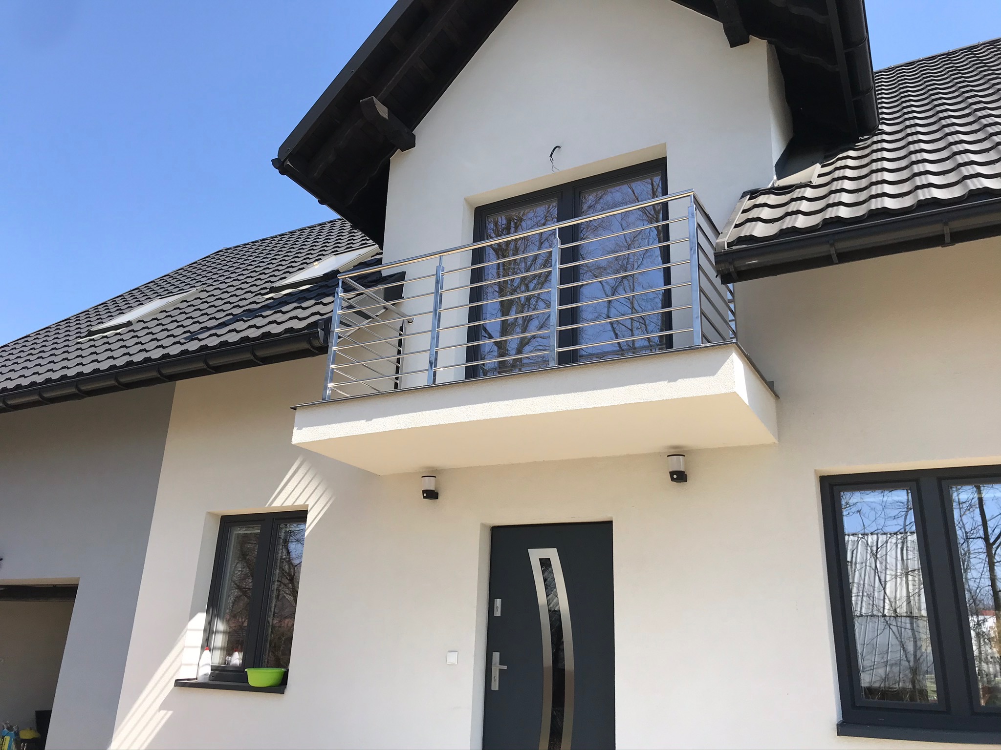 Biały dom z balkonem i balustradą ze stali nierdzewnej, widok z dołu na tle błękitnego nieba.