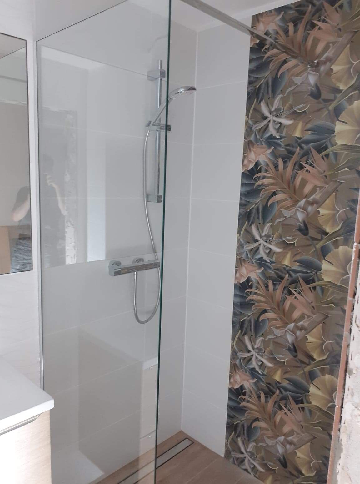 Nowoczesna kabina prysznicowa z białymi płytkami i ścianą akcentową z tapetą w motywy roślinne, widoczny odpływ liniowy w podłodze wyłożonej drewnopodobnymi płytkami.
