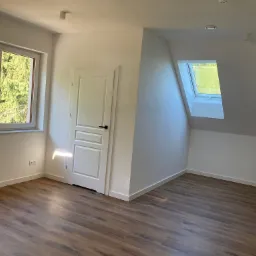 Jasne wnętrze poddasza z drewnopodobnymi panelami na podłodze, białymi ścianami, oknem dachowym i białymi drzwiami z czarną klamką.