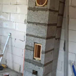 Wznoszenie komina z szarych bloczków betonowych w surowym wnętrzu, widoczne otwory wentylacyjne i rura wbudowana w konstrukcję, poziomica oparta o ścianę, ślady zaprawy na podłodze.