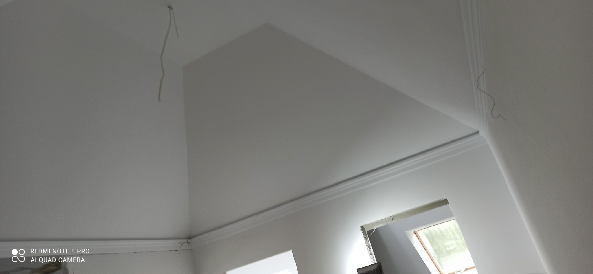 Białe, świeżo pomalowane poddasze z listwami dekoracyjnymi przy suficie, widoczne okno dachowe i wystające kable elektryczne.