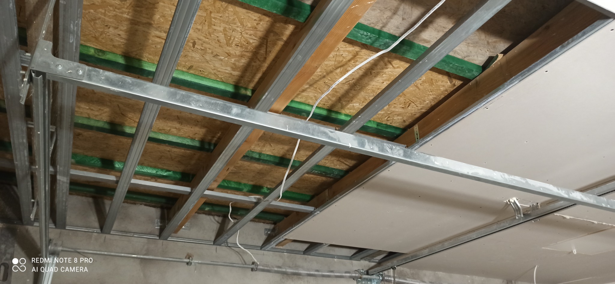 Sufit podwieszany z metalowym stelażem, widoczne płyty OSB z zieloną izolacją, instalacja elektryczna w trakcie montażu.