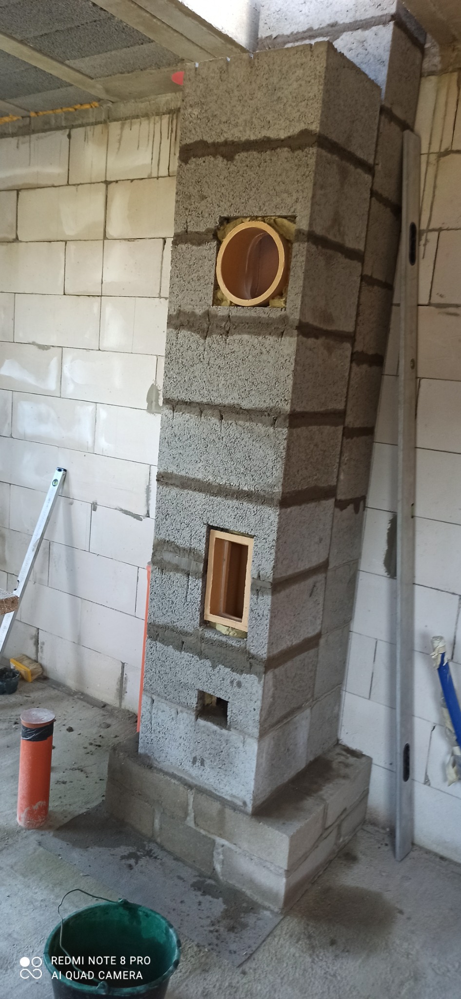Wznoszenie komina z szarych bloczków betonowych w surowym wnętrzu, widoczne otwory wentylacyjne i rura wbudowana w konstrukcję, poziomica oparta o ścianę, ślady zaprawy na podłodze.