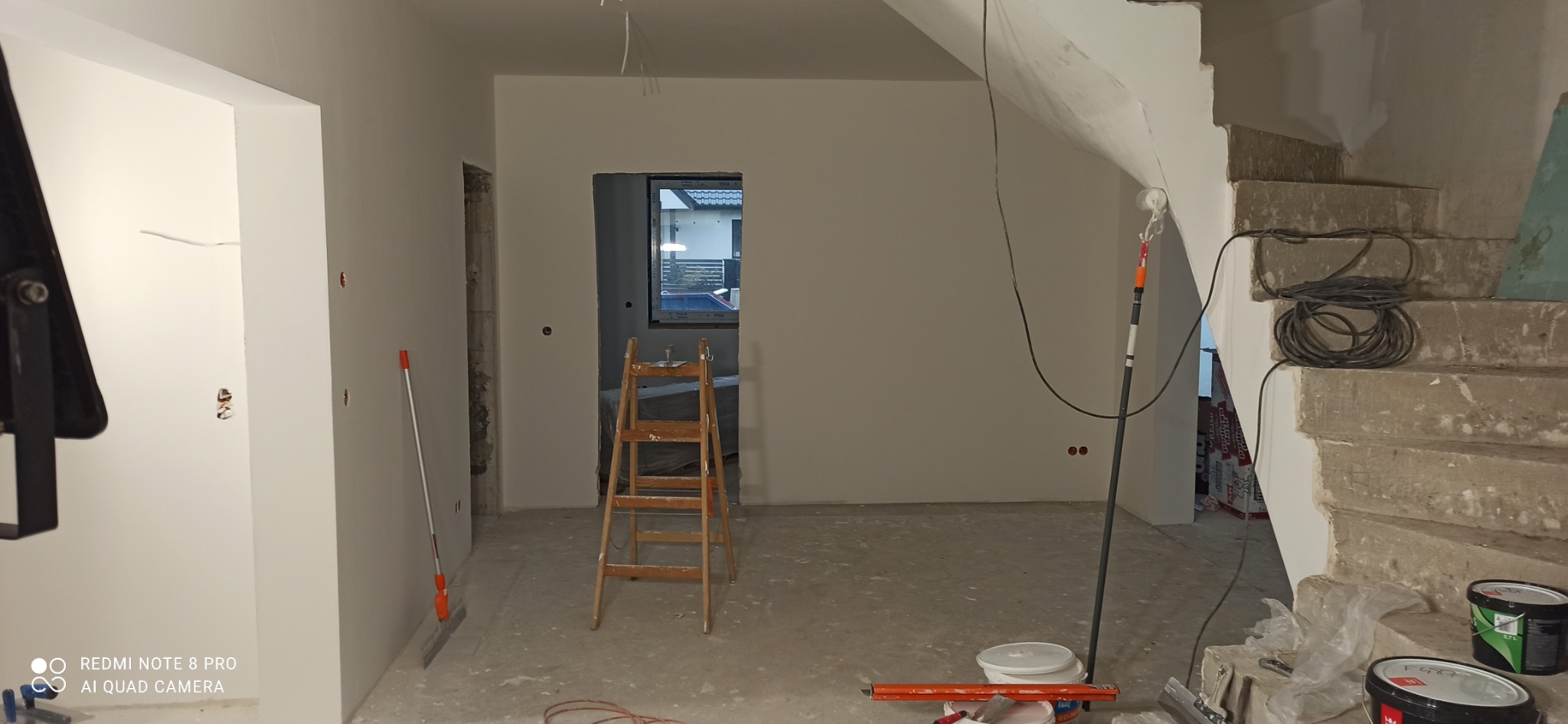 Wnętrze w trakcie remontu: świeżo pomalowane ściany na biało, drewniana drabina, narzędzia malarskie, surowe schody betonowe z widocznymi kablami i opakowaniami materiałów budowlanych.