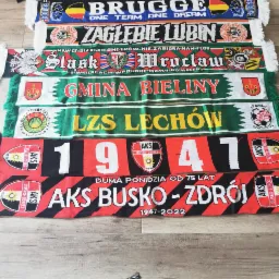 Kolekcja szalików kibicowskich różnych drużyn piłkarskich, ułożonych jeden na drugim, z widocznymi nazwami klubów, herbami i hasłami.