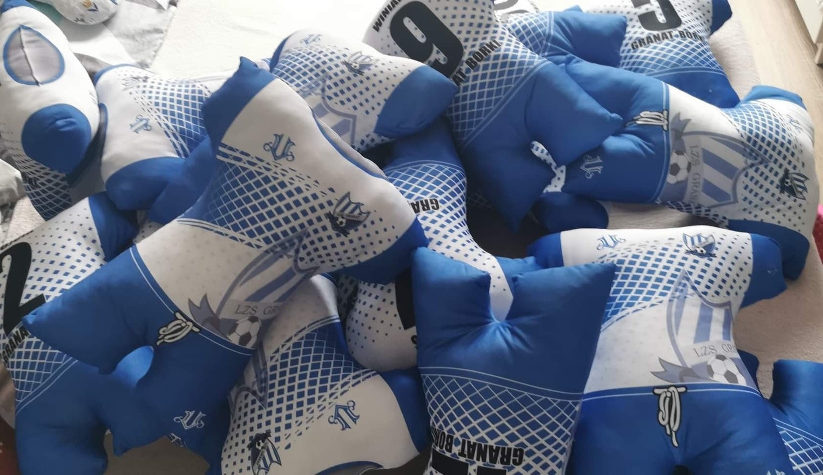 Stos poduszek dekoracyjnych w kształcie koszulek piłkarskich w barwach biało-niebieskich z nadrukowanymi herbami i numerami drużyn sportowych.