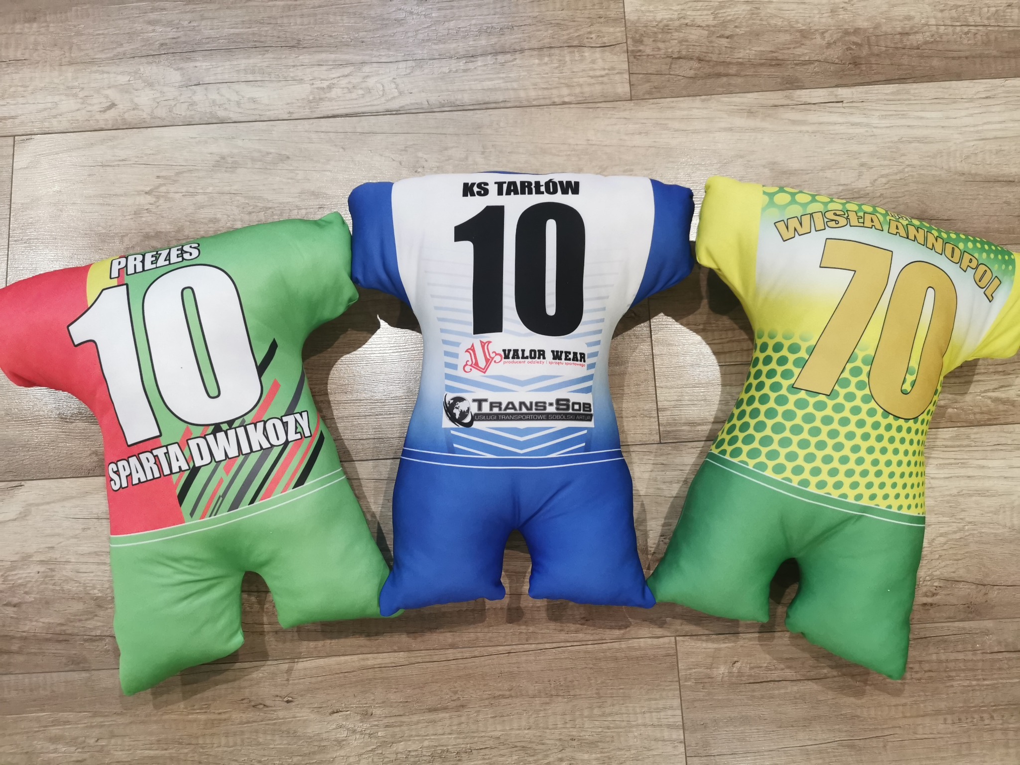 Trzy poduszki w kształcie koszulek sportowych z nadrukami nazw klubów i numerów, leżące na drewnianej podłodze: Prezes 10 Sparta Dwikozy, KS Tarłów 10 Valor Wear, Wisła Annopol 70.