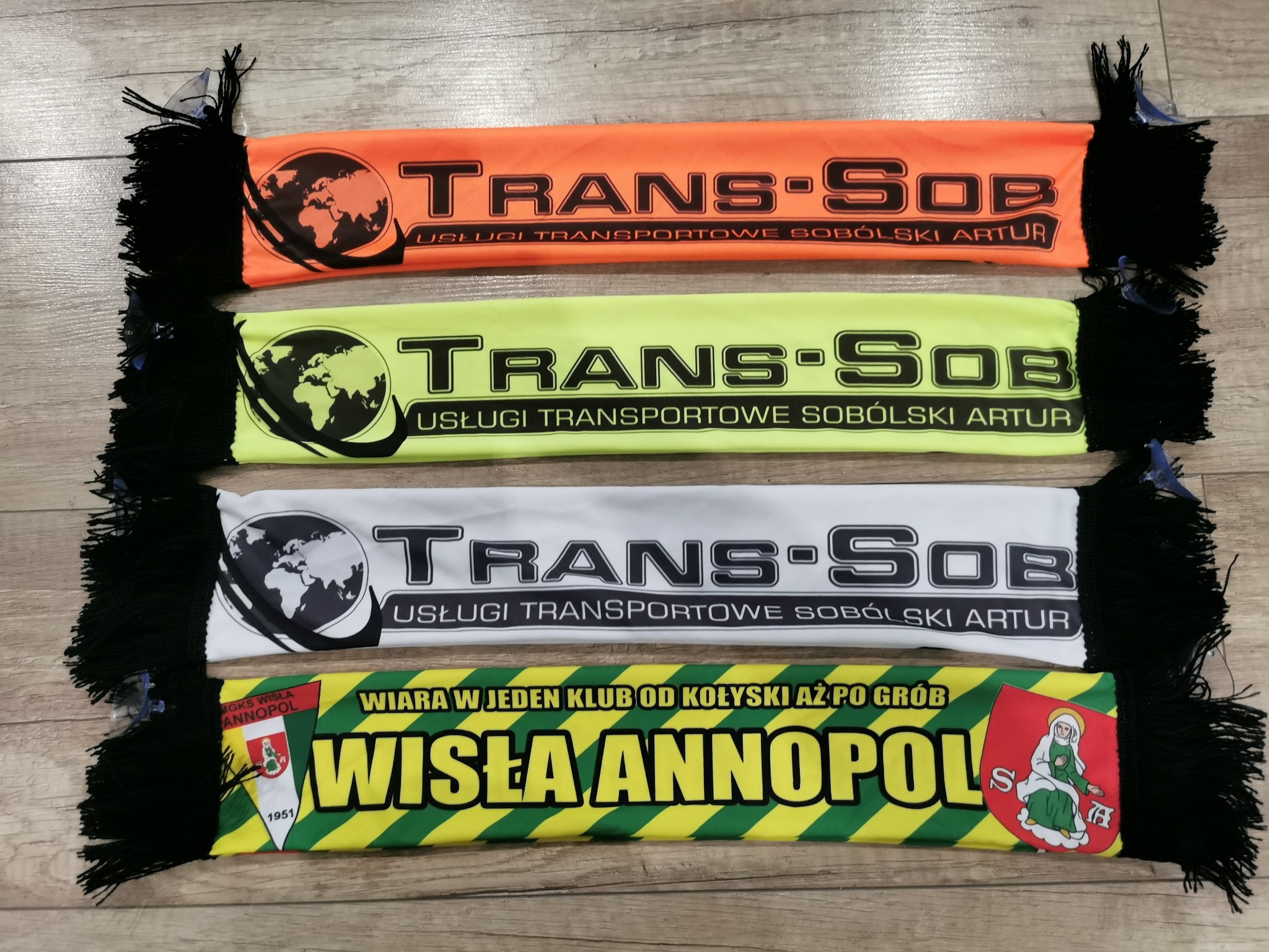 Cztery szaliki z nadrukiem leżące na drewnianej powierzchni. Trzy szaliki z napisem 'TRANS-SOB Usługi Transportowe Sobólski Artur' w kolorach pomarańczowym, żółtym i białym. Czwarty szalik...