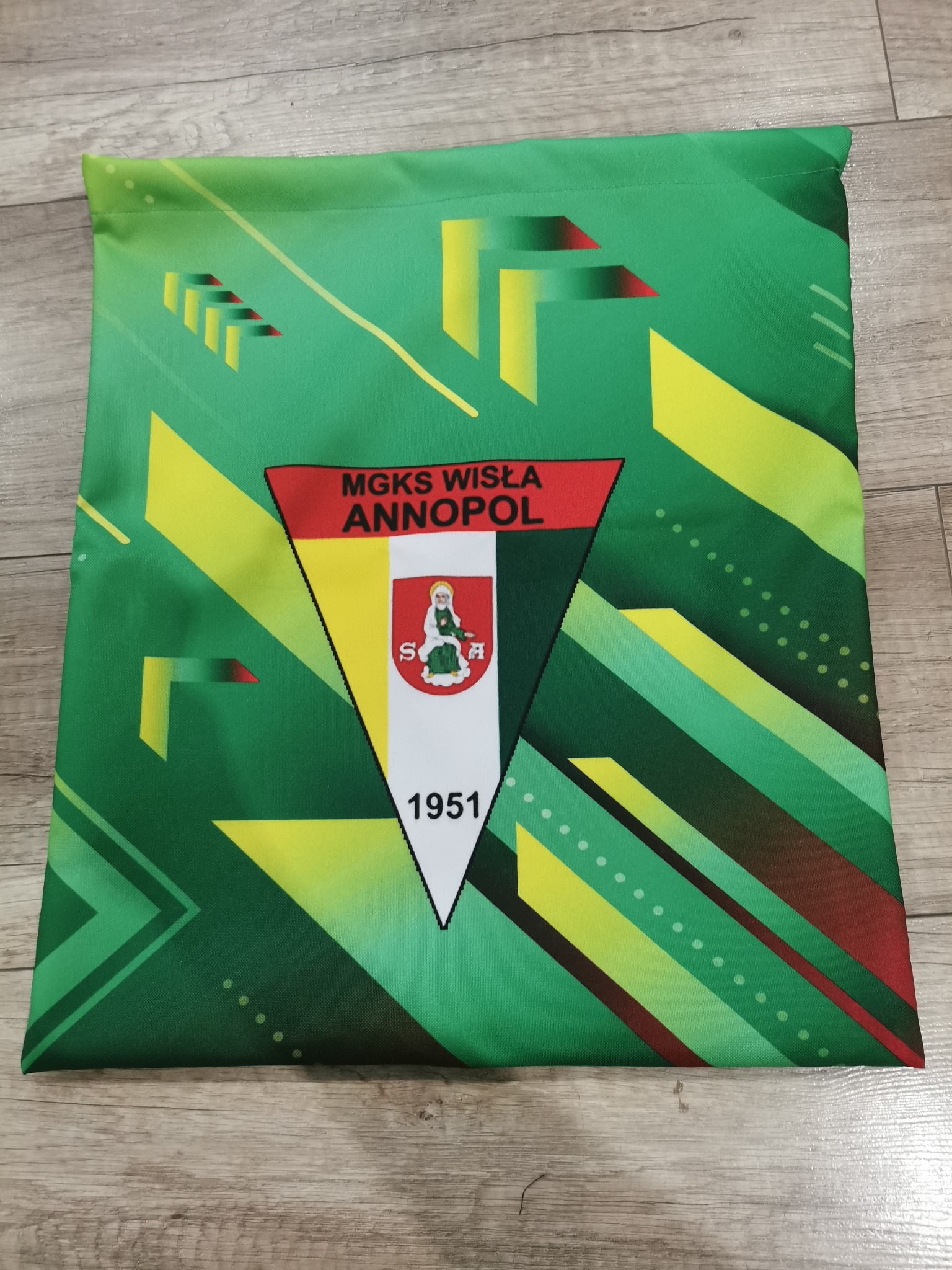 Zielony worek z nadrukowanym logo klubu sportowego MGKS Wisła Annopol z datą 1951, leżący na drewnianej podłodze.
