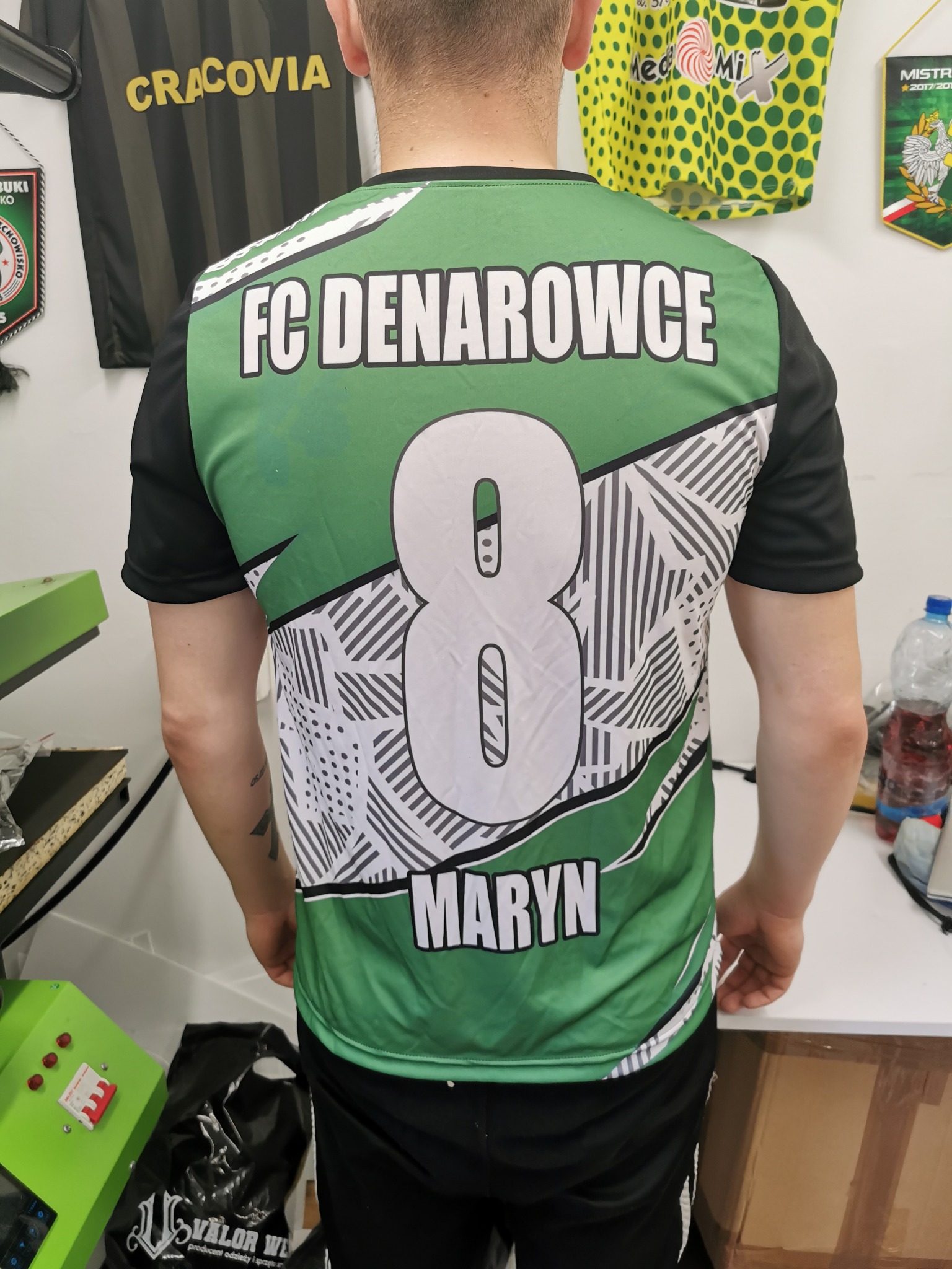 Tył koszulki sportowej z nadrukiem 'FC Denarowce' i numerem 8, wykonanej w Tarłowie, prezentowany na osobie stojącej w pomieszczeniu z innymi koszulkami i akcesoriami.