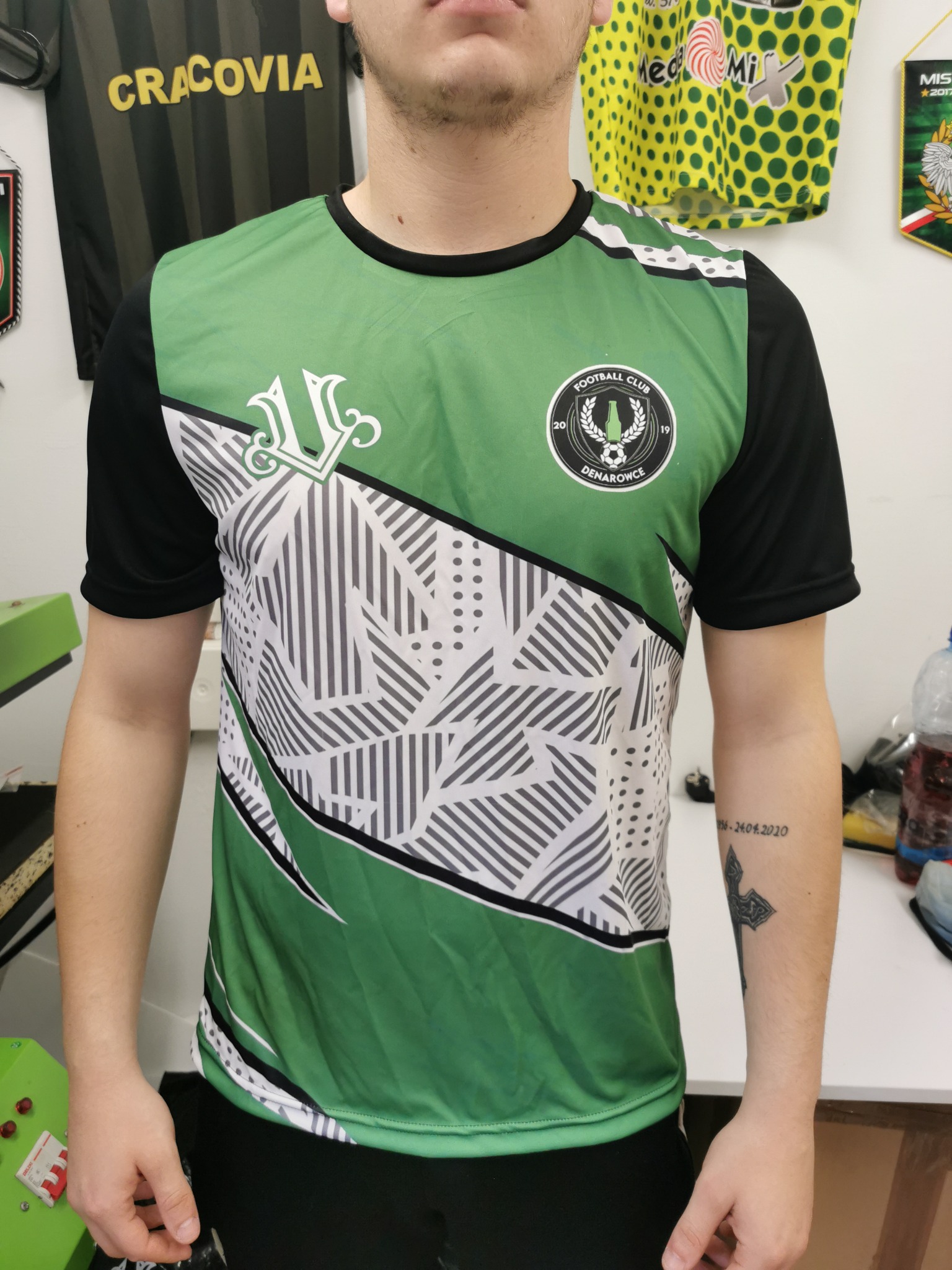 Mężczyzna w zielono-czarnej koszulce sportowej z nadrukiem herbu i nazwy klubu 'Football Club Denarowice', z widocznym tatuażem na przedramieniu.