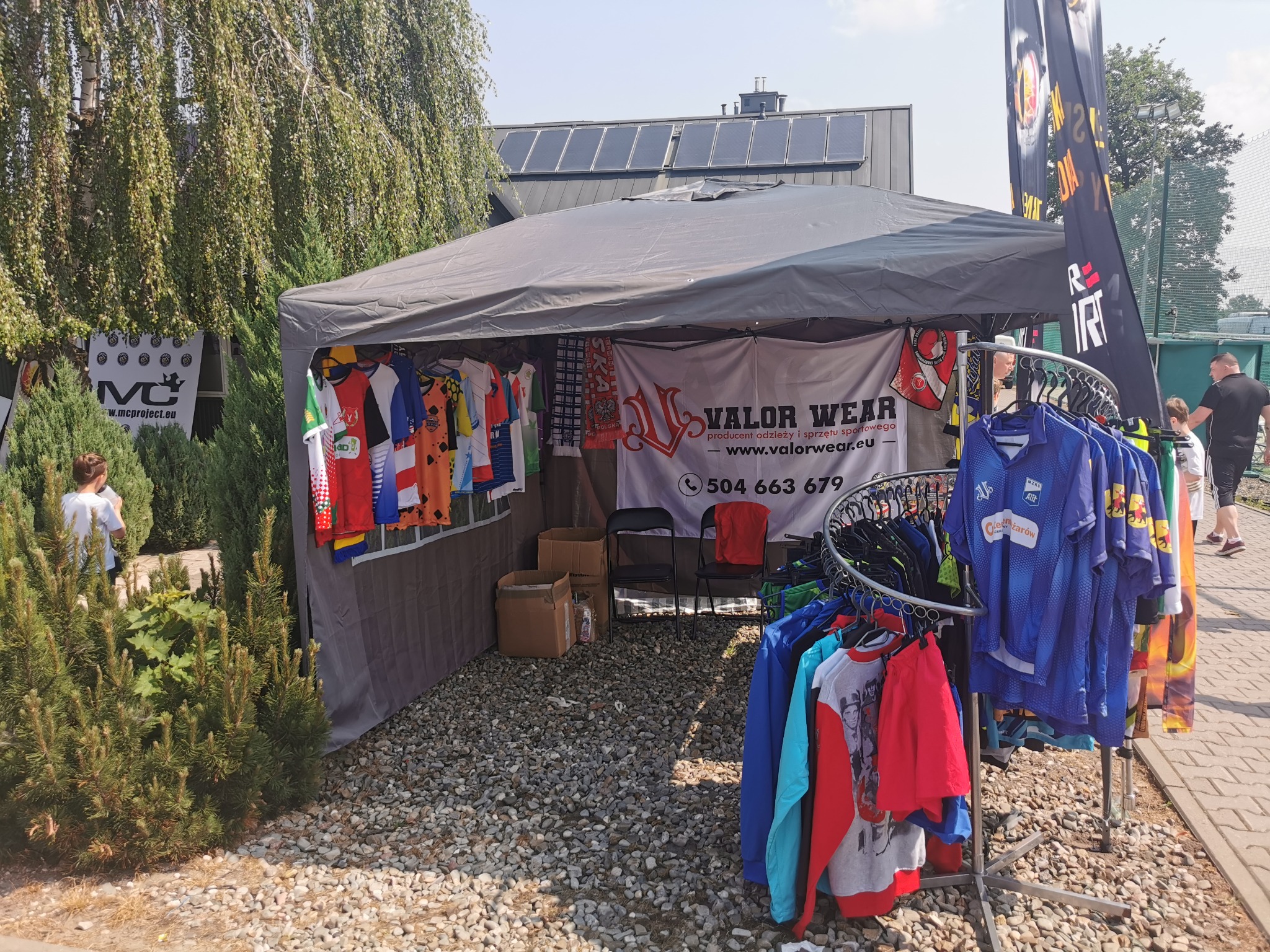 Stoisko handlowe z odzieżą sportową Valor Wear na zewnątrz, z ubraniami na wieszakach, flagami reklamowymi i kartonami w tle, ustawione na żwirowym podłożu.