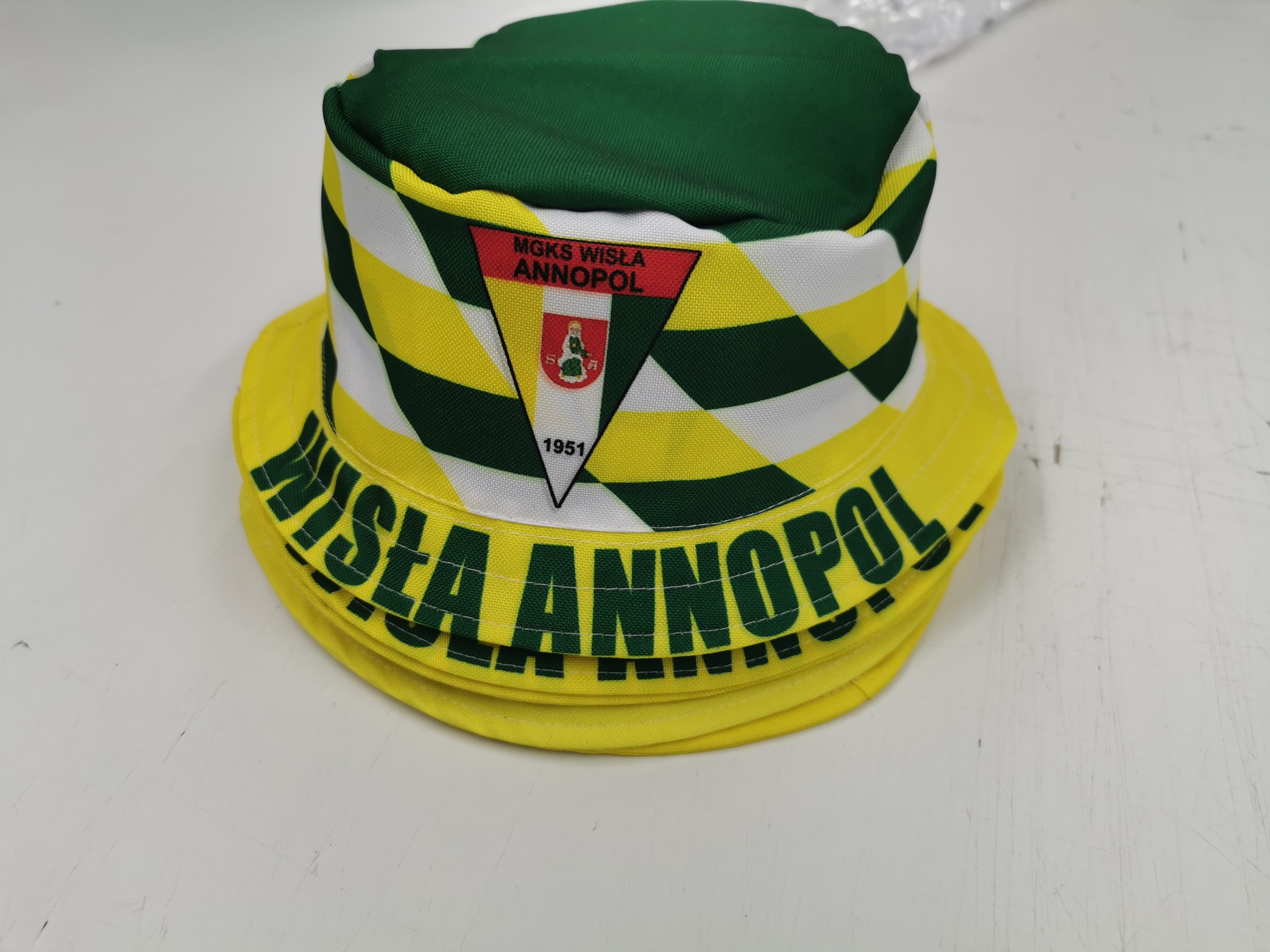 Żółto-zielono-biały kapelusz typu bucket hat z nadrukowanym herbem i napisem 'WISŁA ANNOPOL' na rondzie, leżący na białym tle.