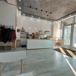 Jasne wnętrze kawiarni z białymi ścianami, drewnianymi akcentami, betonowym sufitem i rzędem wiszących lamp, widok na ladę z pracownikiem i wejście z przeszklonymi drzwiami.