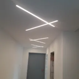 Korytarz z nowoczesnym oświetleniem sufitowym w postaci podłużnych lamp LED ułożonych w geometryczne wzory, szara ściana i drzwi w tle, widoczne materiały budowlane na podłodze.