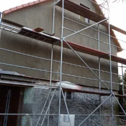 Dom w trakcie prac elewacyjnych, widoczne rusztowanie, świeżo nałożony tynk na górnej części ściany, surowa dolna część z widoczną strukturą kamienia, okno z folią zabezpieczającą.