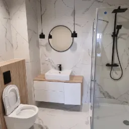 Nowoczesna łazienka w Gorzowie Wielkopolskim z marmurowymi płytkami, podwieszaną toaletą z drewnianą obudową, okrągłym lustrem z czarną ramą, białą szafką z drewnianym blatem i kabiną prysznicową...