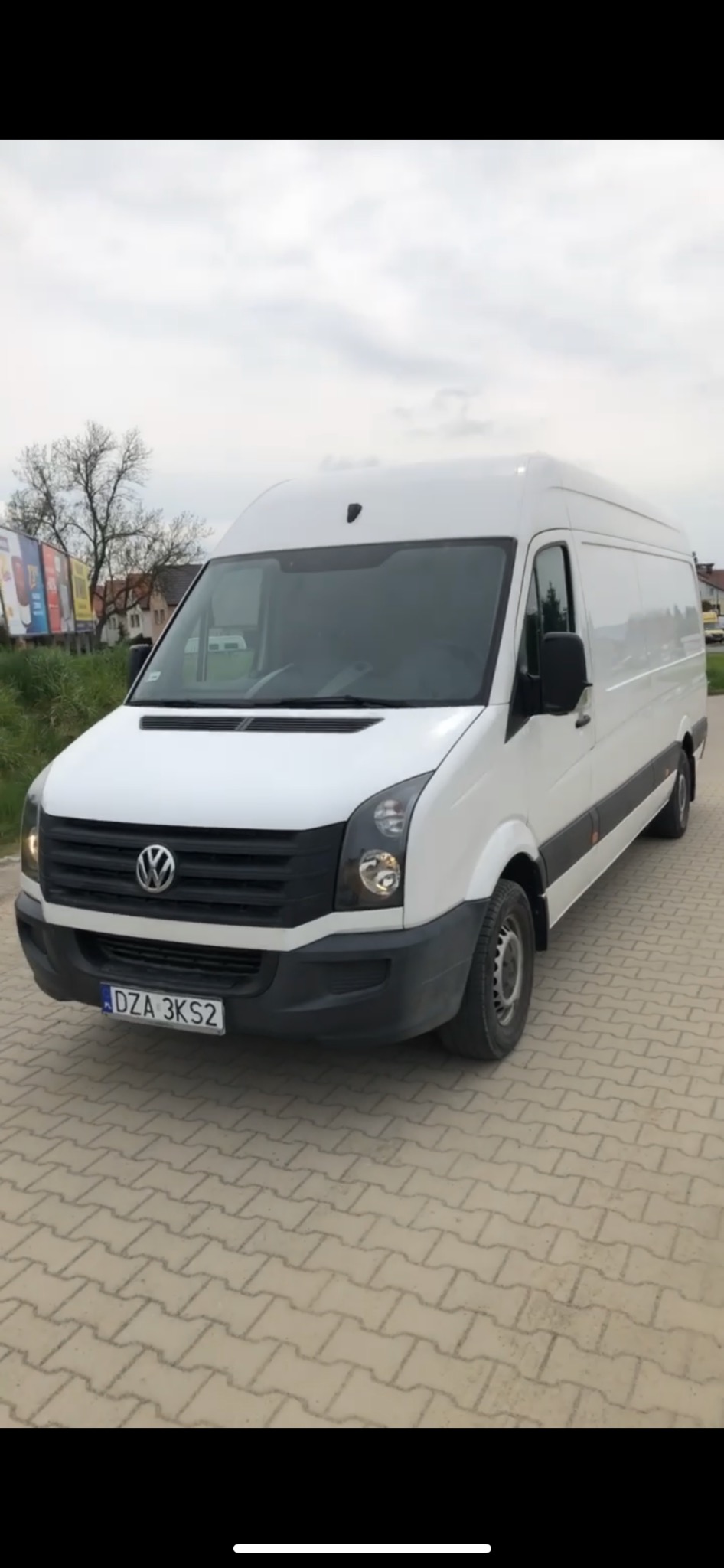 Biały samochód dostawczy marki Volkswagen Crafter zaparkowany na brukowanym placu, z widoczną tablicą rejestracyjną DZA 3KS2 i drzewami w tle.