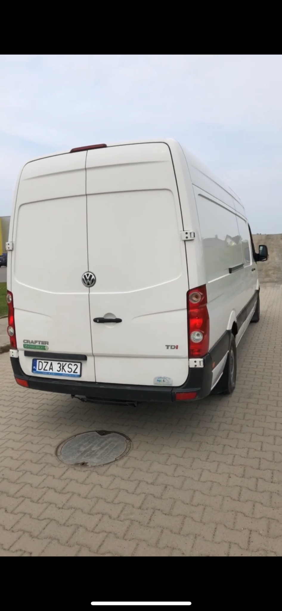 Tył białego samochodu dostawczego Volkswagen Crafter TDI z widoczną tablicą rejestracyjną DZA 3KS2, zaparkowanego na chodniku z kostki brukowej, z deklem studzienki kanalizacyjnej w tle.