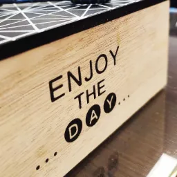 Drewniane pudełko z napisem 'Enjoy the day...' i wzorem geometrycznym na górze, na którym leżą nożyczki fryzjerskie z niebieskimi uchwytami.