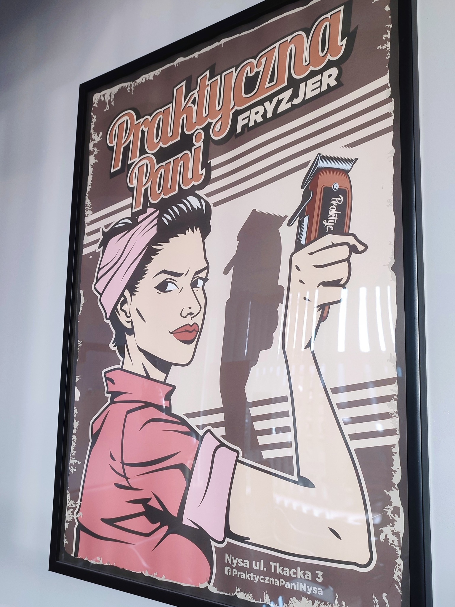 Oprawiony plakat w stylu retro przedstawiający kobietę z podwiniętymi rękawami i bandaną, trzymającą maszynkę do strzyżenia; napis 'Praktyczna Pani' i adres 'Nysa ul. Tkacka 3'.