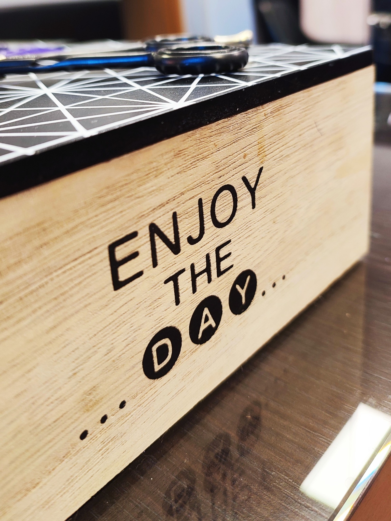 Drewniane pudełko z napisem 'Enjoy the day...' i wzorem geometrycznym na górze, na którym leżą nożyczki fryzjerskie z niebieskimi uchwytami.