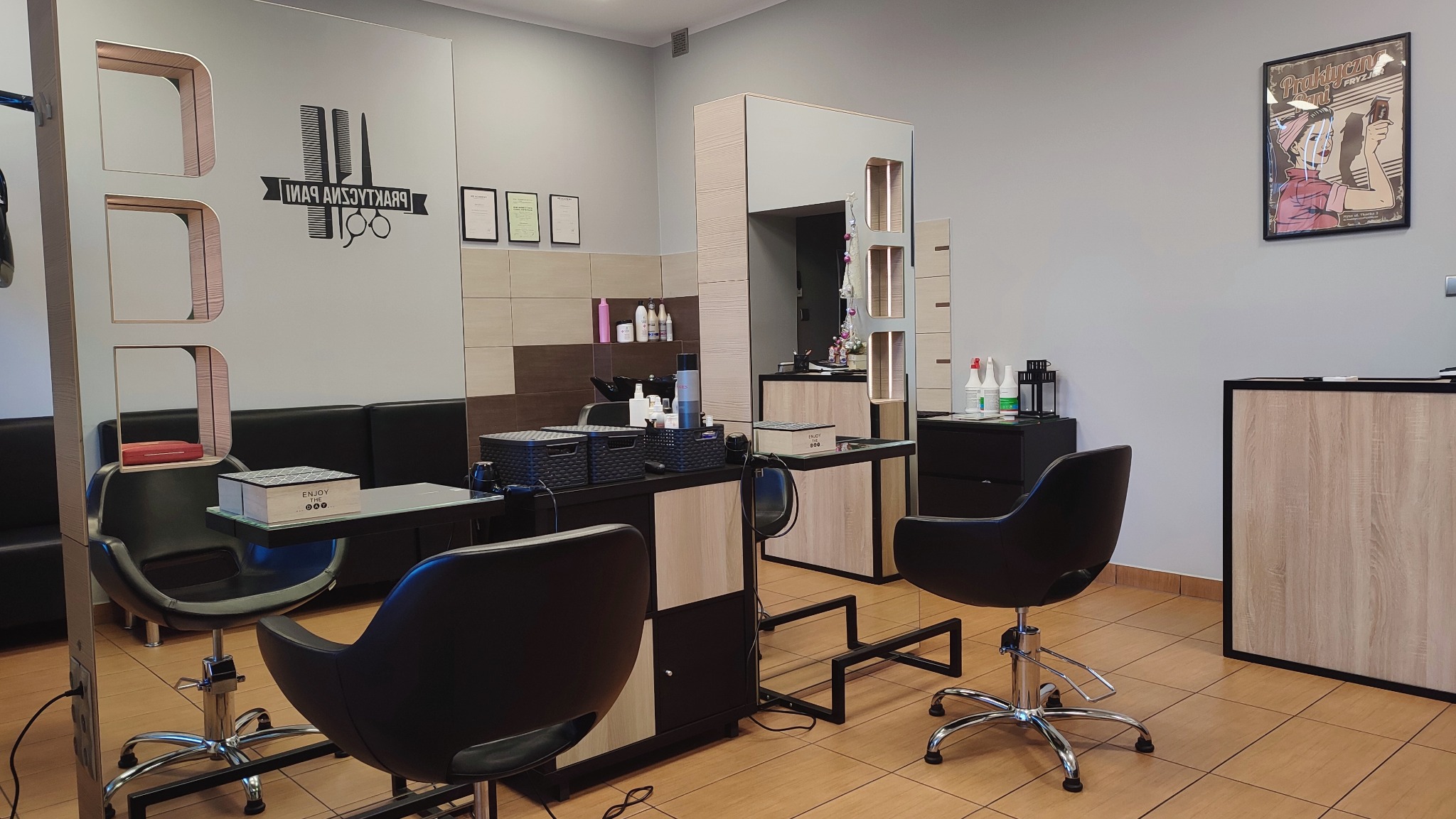 Wnętrze salonu z dwoma stanowiskami, czarnymi fotelami na srebrnych podstawach, lustrami i drewnianymi elementami wykończenia, na ścianie grafika z logo salonu.