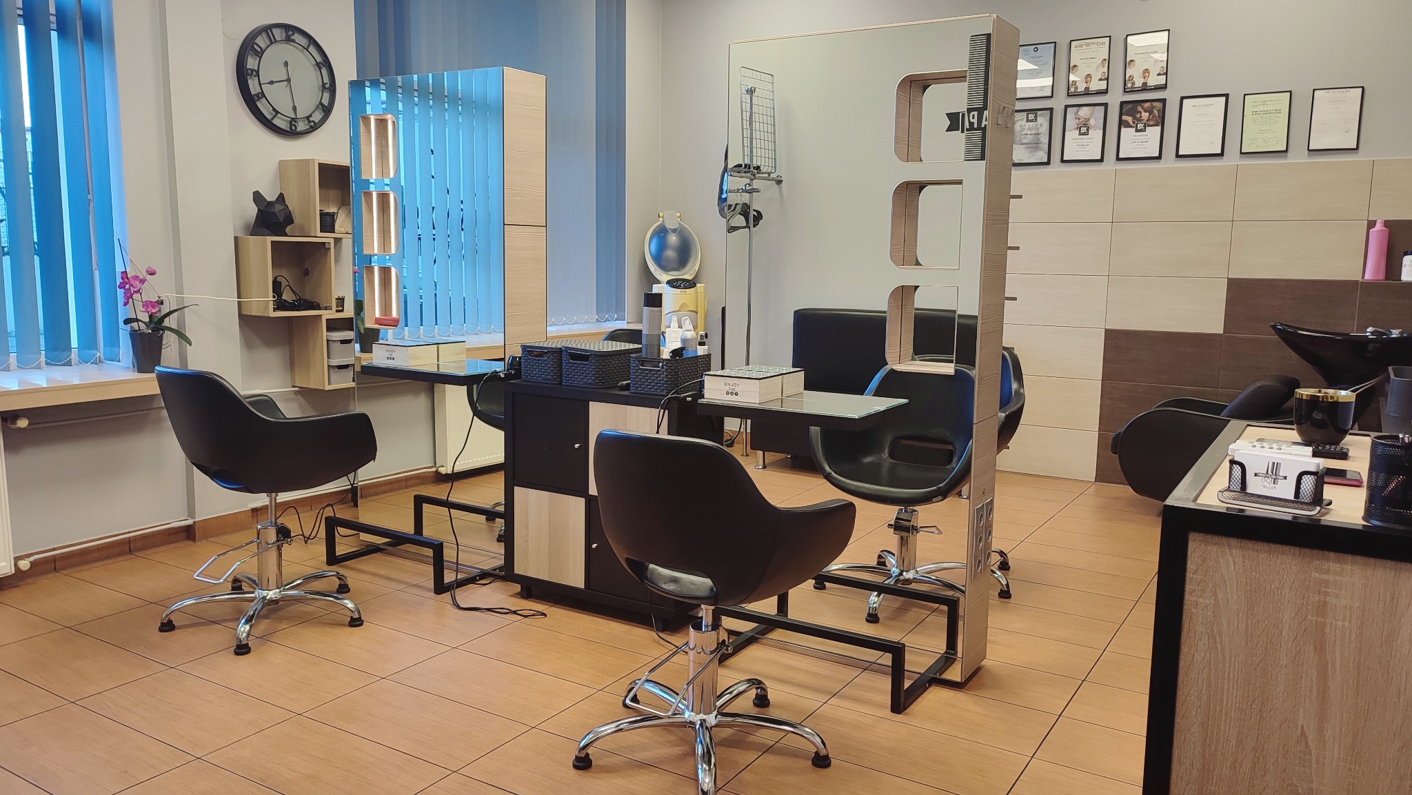 Wnętrze salonu z trzema stanowiskami: czarne fotele na chromowanych podstawach, lustra z oświetleniem, szafki z akcesoriami, dyplomy na ścianie i zegar.