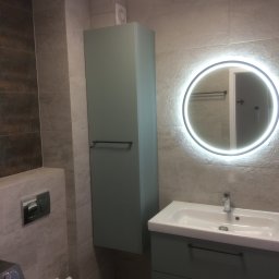 Gres Andrzej - Nowoczesna łazienka z szarymi płytkami, podświetlanym okrągłym lustrem, wiszącą szafką i umywalką z szufladami.