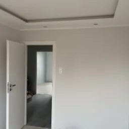 Jasne wnętrze w trakcie odświeżania: pomalowane ściany na szaro, otwarte białe drzwi, widoczna podłoga imitująca drewno i fragment sufitu z dekoracyjnym obramowaniem.