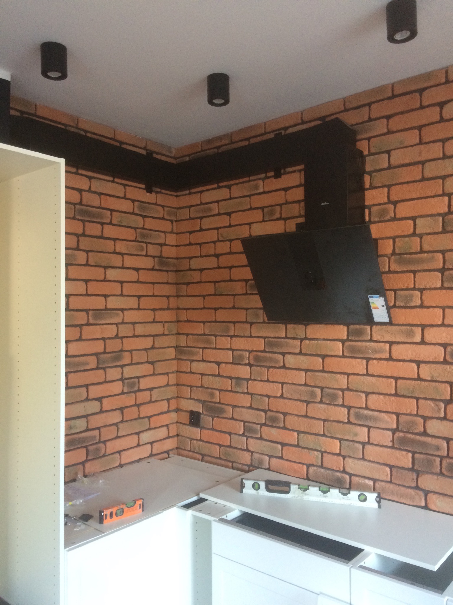 Fragment kuchni w trakcie montażu, widoczna ściana z cegły, czarny okap Amica, białe szafki i poziomice na blacie.
