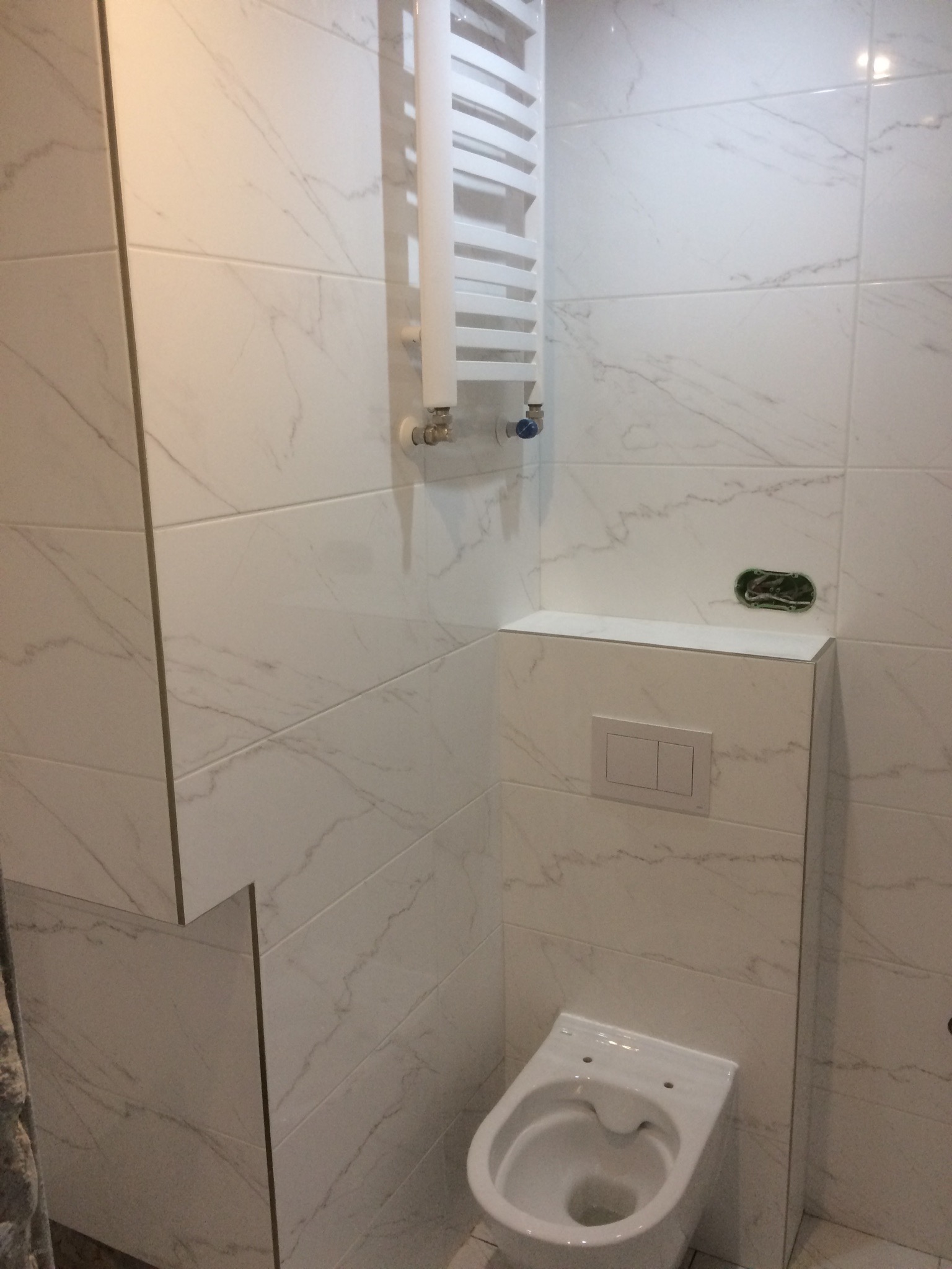 Nowoczesna łazienka z białymi płytkami imitującymi marmur, wisząca toaleta z zabudową, grzejnik drabinkowy i widoczne przygotowanie do podłączenia oświetlenia.